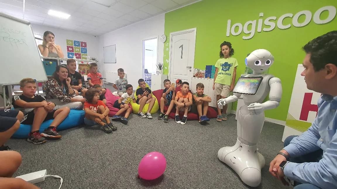 Copii așezați pe podea ascultă robotul Pepper într-o sală de curs